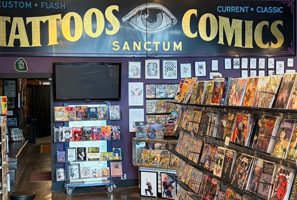 Interior of a shop with comic books displayed on racks, tattoo art on the walls, and a sign above reading "Tattoos Sanctum Comics" in large gold letters, giving the salon an eclectic and creative atmosphere.