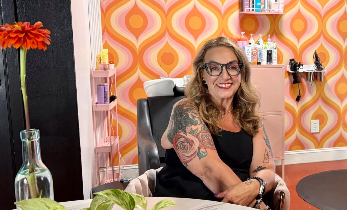 A woman with glasses and tattoos sits smiling in a retro-themed salon, Sanctum, where geometric orange wallpaper and shelves of hair products create a vibrant backdrop.