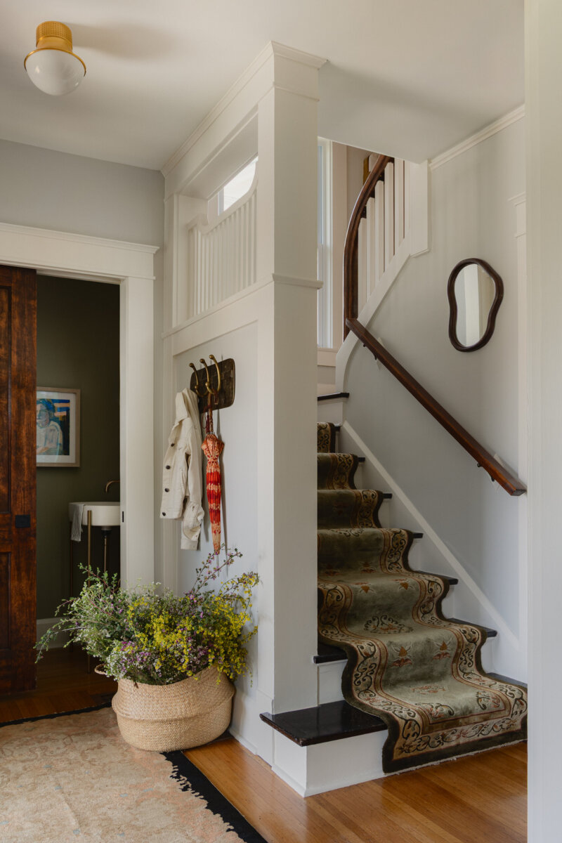 A small entryway in this historic bungalow exudes preserved charm, featuring a carpeted staircase, wall hooks holding coats and an umbrella, a mirror, and a woven basket filled with flowers resting on the wooden floor.
