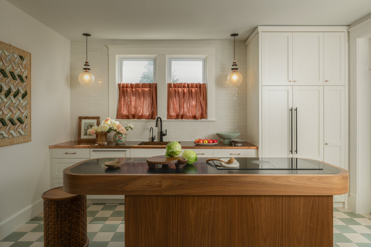 Modern kitchen with a wooden island, green and white checkered floor, white cabinets, globe pendant lights, and orange curtains on two windows above the sink.