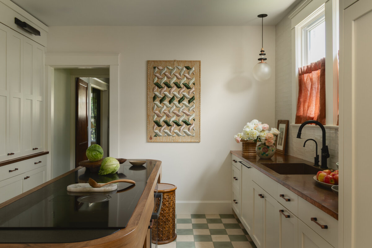 A tidy kitchen with white cabinets, a wood countertop, a checkered floor, a kitchen island, and a window with a red curtain exudes the preserved charm of a historic bungalow. A woven wall hanging decorates the wall above a flower vase, adding an art-filled touch to the inviting space.