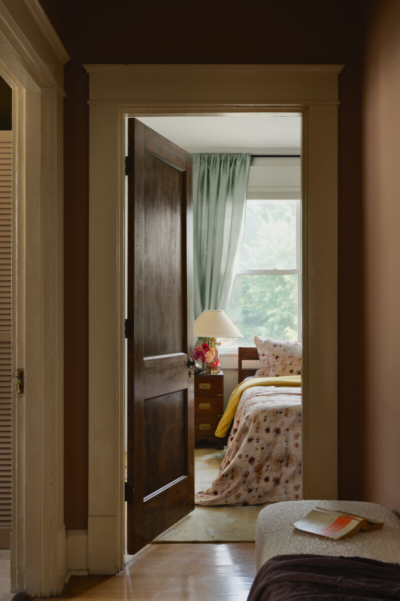 View through a doorway into a sunlit bedroom with floral bedding, a nightstand, lamp, and green curtains reflects the art-filled character of this historic bungalow. An open book rests on a bench in the hallway, enhancing the home’s preserved charm.