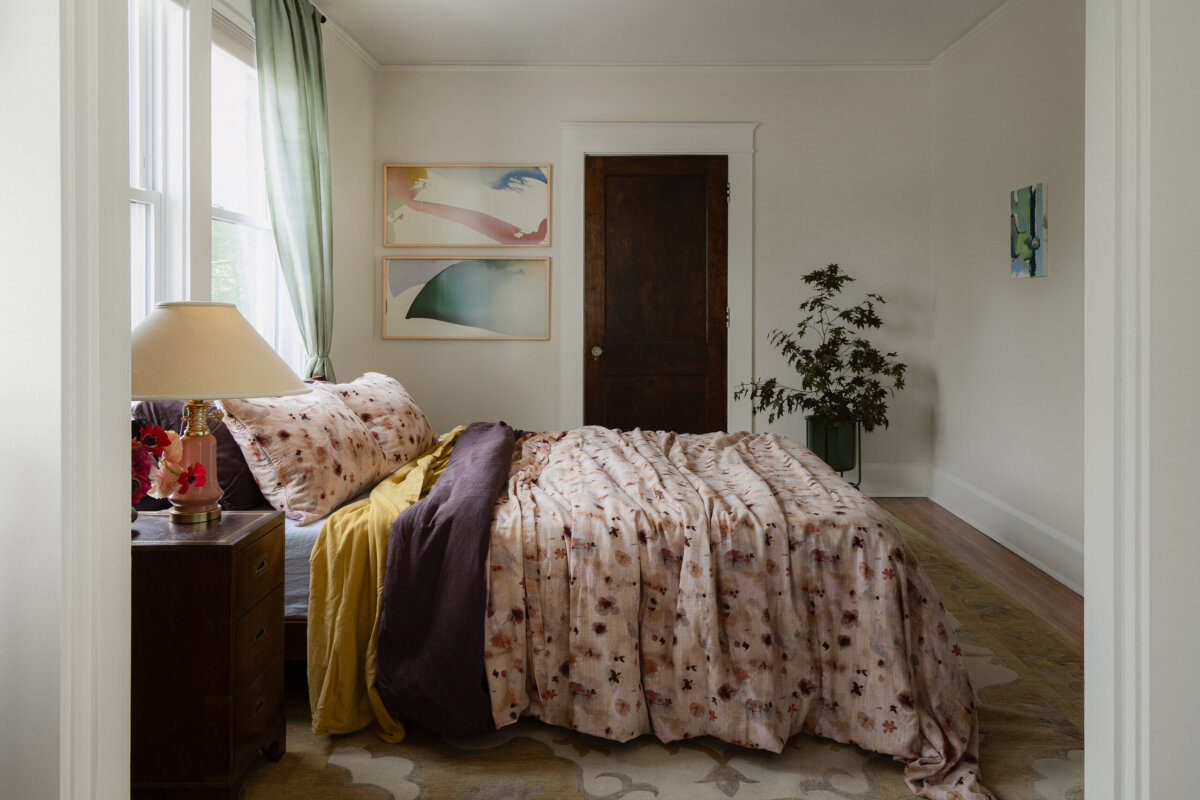 A bedroom in this historic bungalow showcases preserved charm, featuring a double bed with floral bedding, a nightstand with a lamp, art-filled walls with framed pieces above the bed, a potted plant, and a window dressed in sheer green curtains.