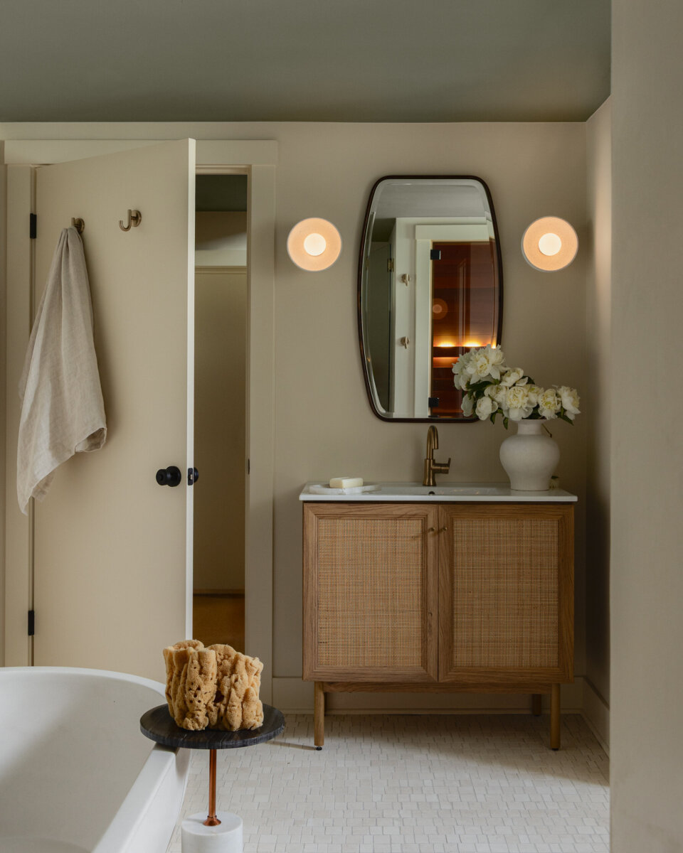 Modern bathroom in an art-filled historic bungalow featuring a vanity, oval mirror, wall lights, white vase with flowers, open door, towel on hook, and bath sponges on a tray near the bathtub.