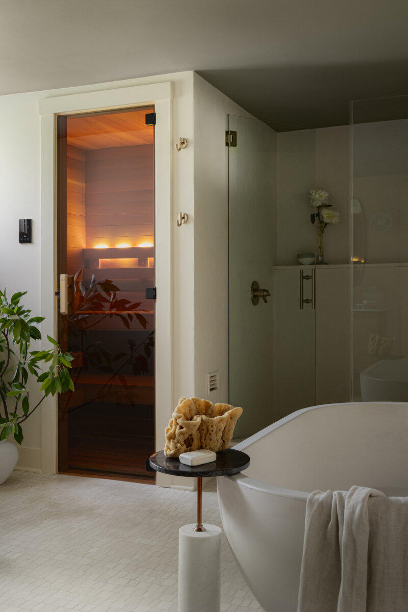 Modern bathroom with a freestanding tub, small side table, plant, and a glass-door sauna softly lit with warm lights in the background, blending preserved charm from a historic bungalow with art-filled accents throughout.
