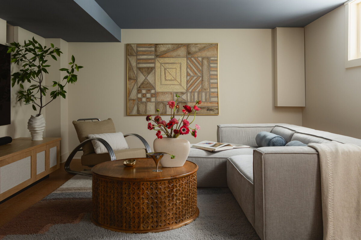 Art-filled and inviting, this modern living room combines a gray sectional sofa, round wooden coffee table, armchair, potted plant, and abstract wall art—creating a perfect blend of contemporary style with the preserved charm of a historic bungalow. Sunlight pours in through the window, illuminating every thoughtfully curated detail.