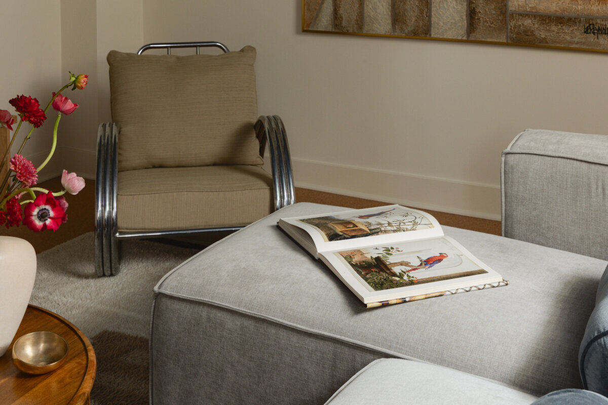 A gray ottoman with an open art book sits next to a beige armchair, adding to the art-filled atmosphere; a vase of red flowers is on a side table, highlighting the preserved charm of this historic bungalow.