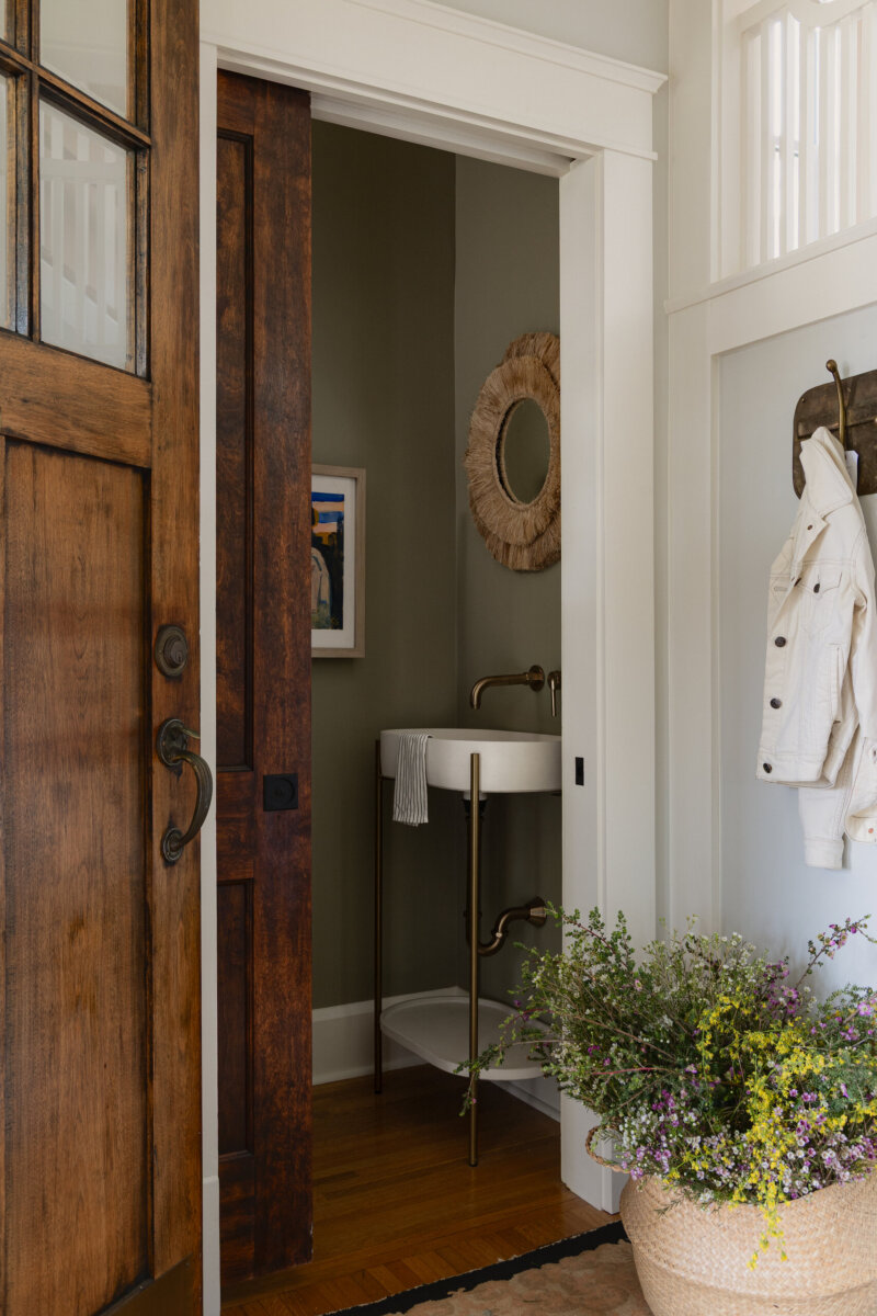 A small bathroom with a pedestal sink, round mirror, wall art, and green walls is visible through a wooden sliding door near an entryway with a hanging white jacket and a vase of flowers, reflecting the preserved charm of this art-filled historic bungalow.