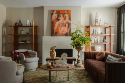 A modern living room with a brown sofa, two armchairs, a round coffee table with sculptures, built-in shelves, large plant, and abstract art above a white-tiled fireplace.