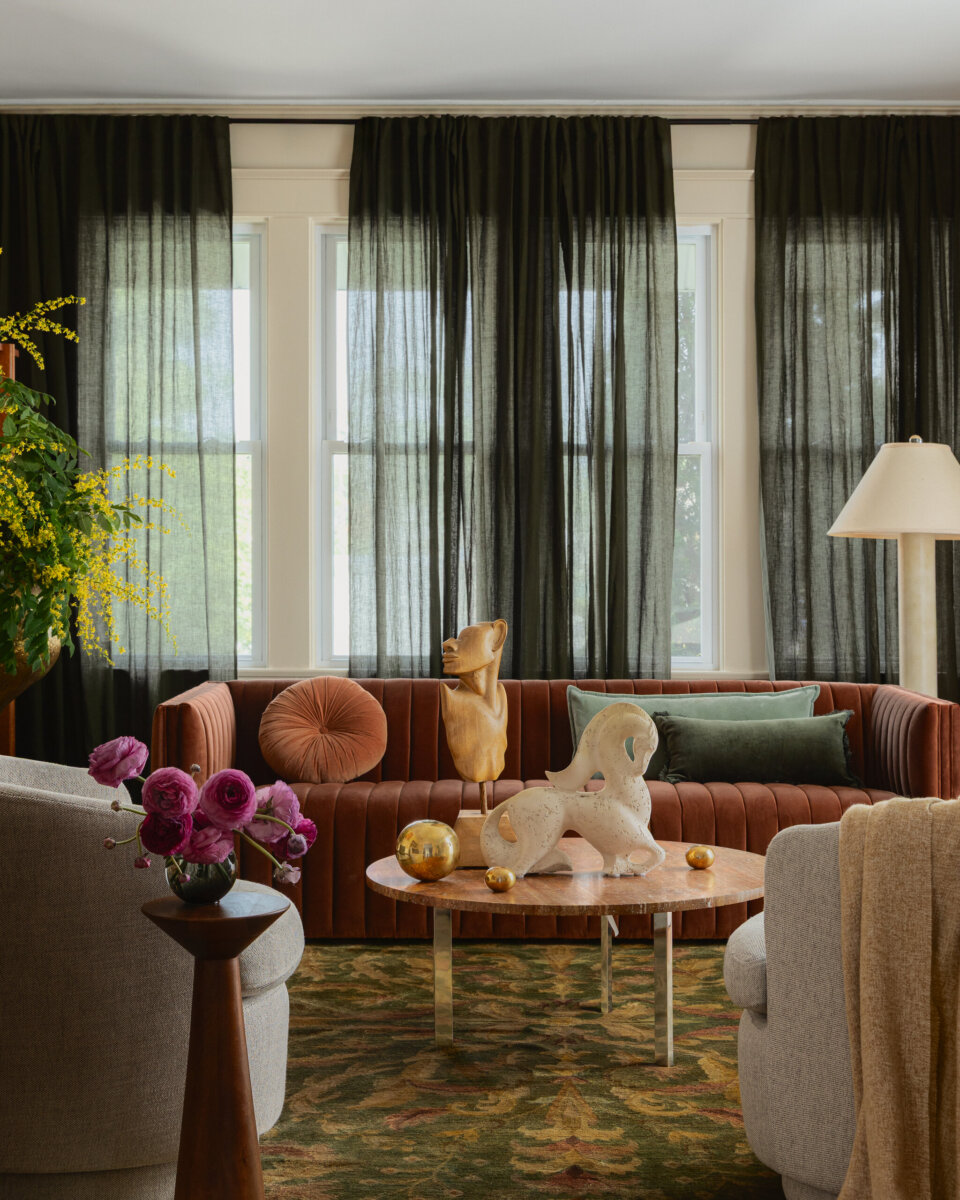A living room with a brown sofa, a round coffee table with sculptures, sheer black curtains, a floor lamp, and flowers in vases beautifully showcases the preserved charm of this art-filled historic bungalow.
