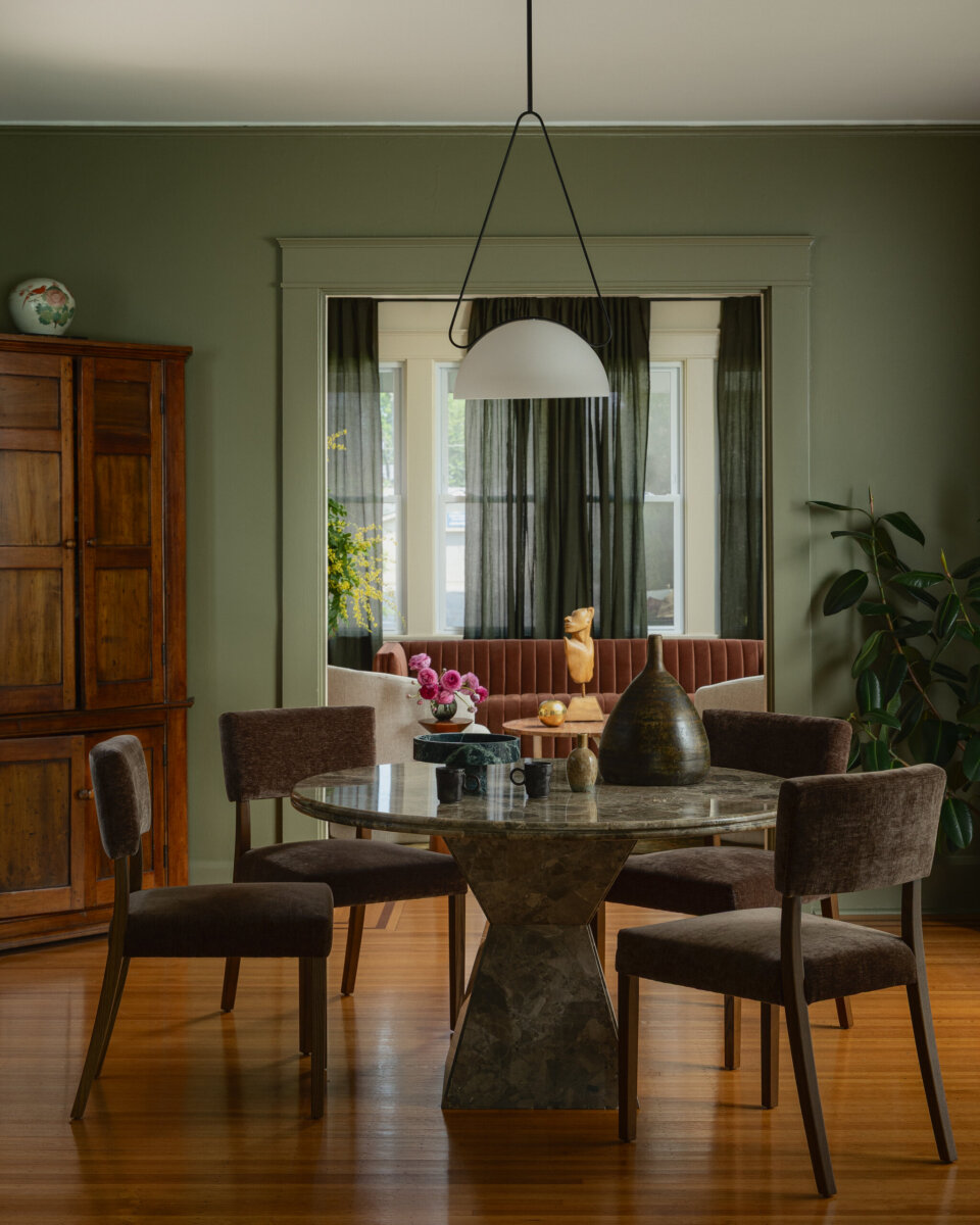 Dining area with a round marble table, four brown chairs, wooden cabinet, potted plant, and pendant light—an art-filled space that offers a view into a sitting room with windows and dark curtains. 