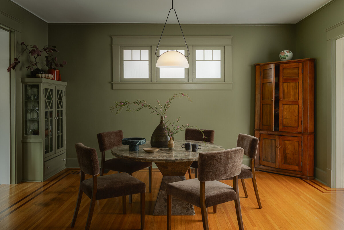 A dining room with a round table, five chairs, a hanging light, wooden cabinets, and a vase with branches on the table. The green walls and hardwood floor reflect the preserved charm of this historic bungalow while creating an inviting atmosphere.