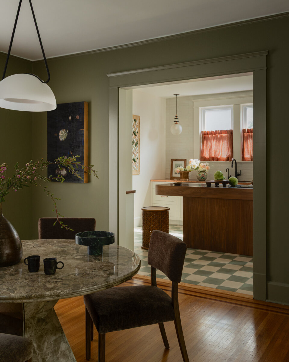 A dining area with a round marble table leads into a kitchen with checkered flooring, an island, and red curtains above two windows, perfectly showcasing the preserved charm of this art-filled historic bungalow.