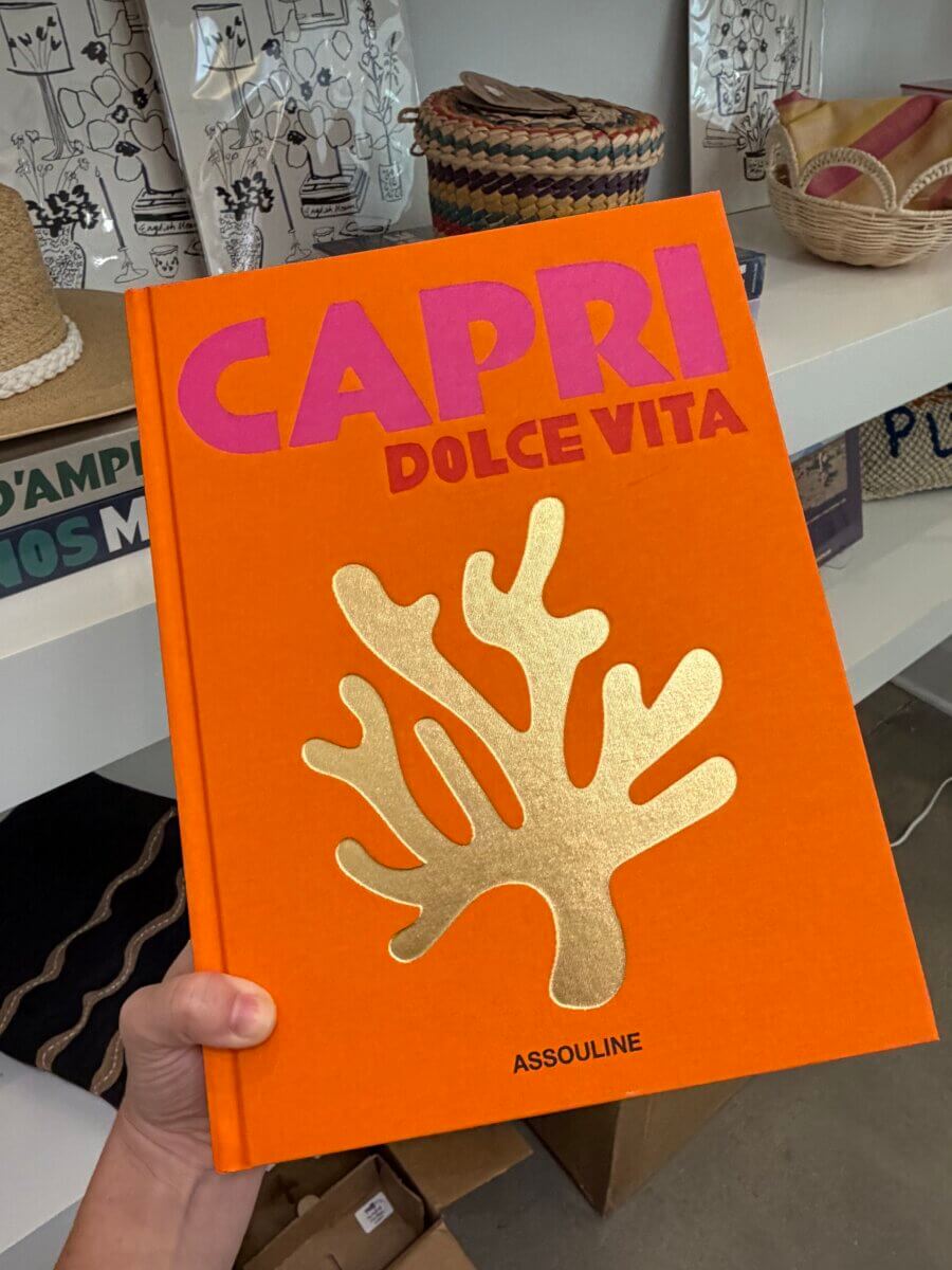 15 Birmingham FINDS for August: Breezy Fashion, Entertaining & More - 16 A hand holds the book "Capri Dolce Vita" with an orange cover and gold coral design, published by Assouline, in front of a shelf displaying Birmingham items such as woven baskets and other unique August finds.