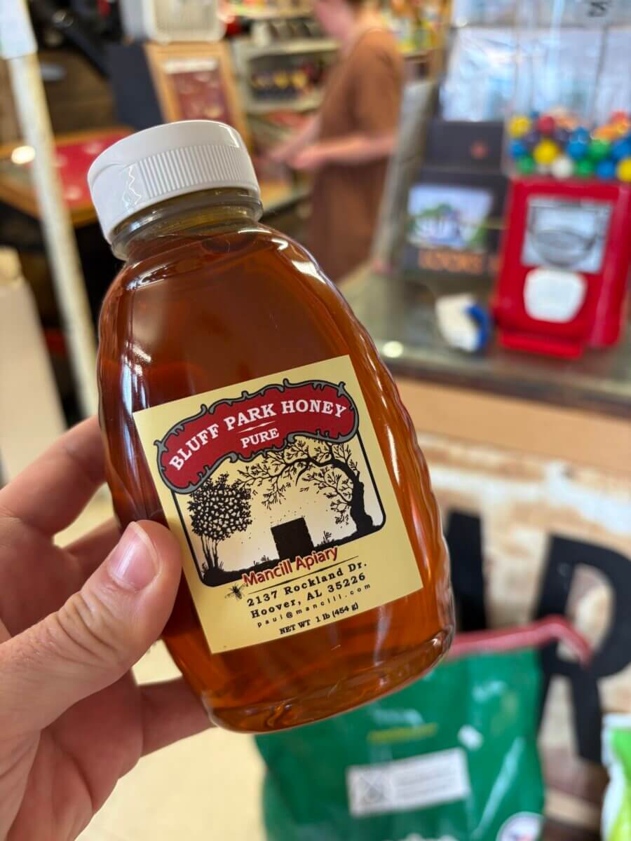 15 Birmingham FINDS for August: Breezy Fashion, Entertaining & More - 4 A hand holds a bottle of Bluff Park Honey from Mancill Apiary inside a Birmingham store, showcasing one of the best local finds this August; in the background, a blurred person and other items are visible.