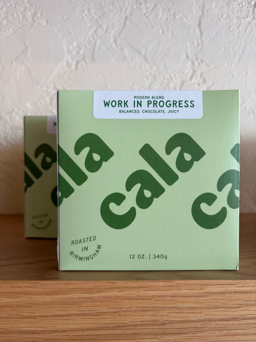 15 Birmingham FINDS for August: Breezy Fashion, Entertaining & More - 9 Light green box of Cala coffee labeled "Work in Progress," listing flavors as balanced, chocolate, juicy. This local find from Birmingham comes in a 12 oz (340g) box and sits on a wooden surface—perfect for savoring on an August morning.