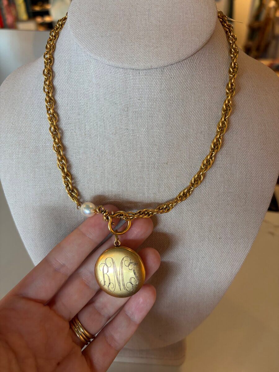 15 Birmingham FINDS for August: Breezy Fashion, Entertaining & More - 6 A hand holds a gold locket with an engraved script on a gold chain necklace, one of the exquisite items displayed on a jewelry bust in Birmingham this August.