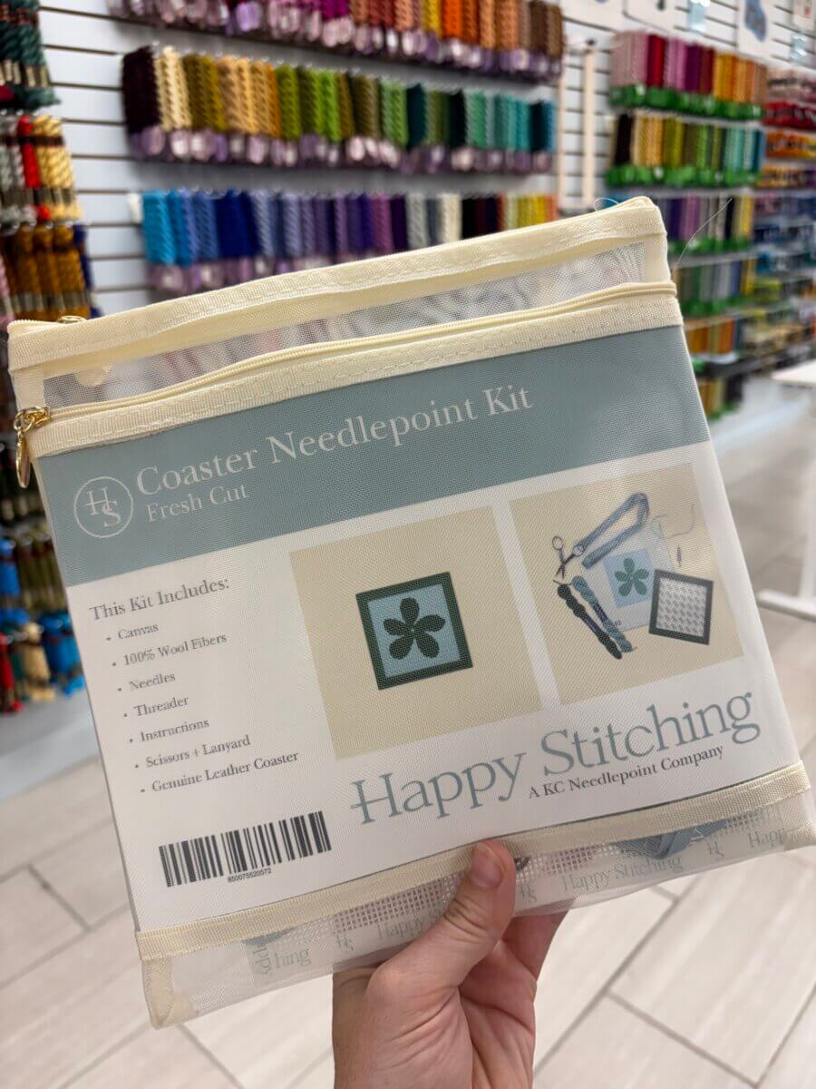 15 Birmingham FINDS for August: Breezy Fashion, Entertaining & More - 13 A hand holds a clear pouch containing a coaster needlepoint kit with supplies, in a Birmingham store with colorful thread on the wall in the background—perfect for your next creative project featured in August Birmingham FINDS.