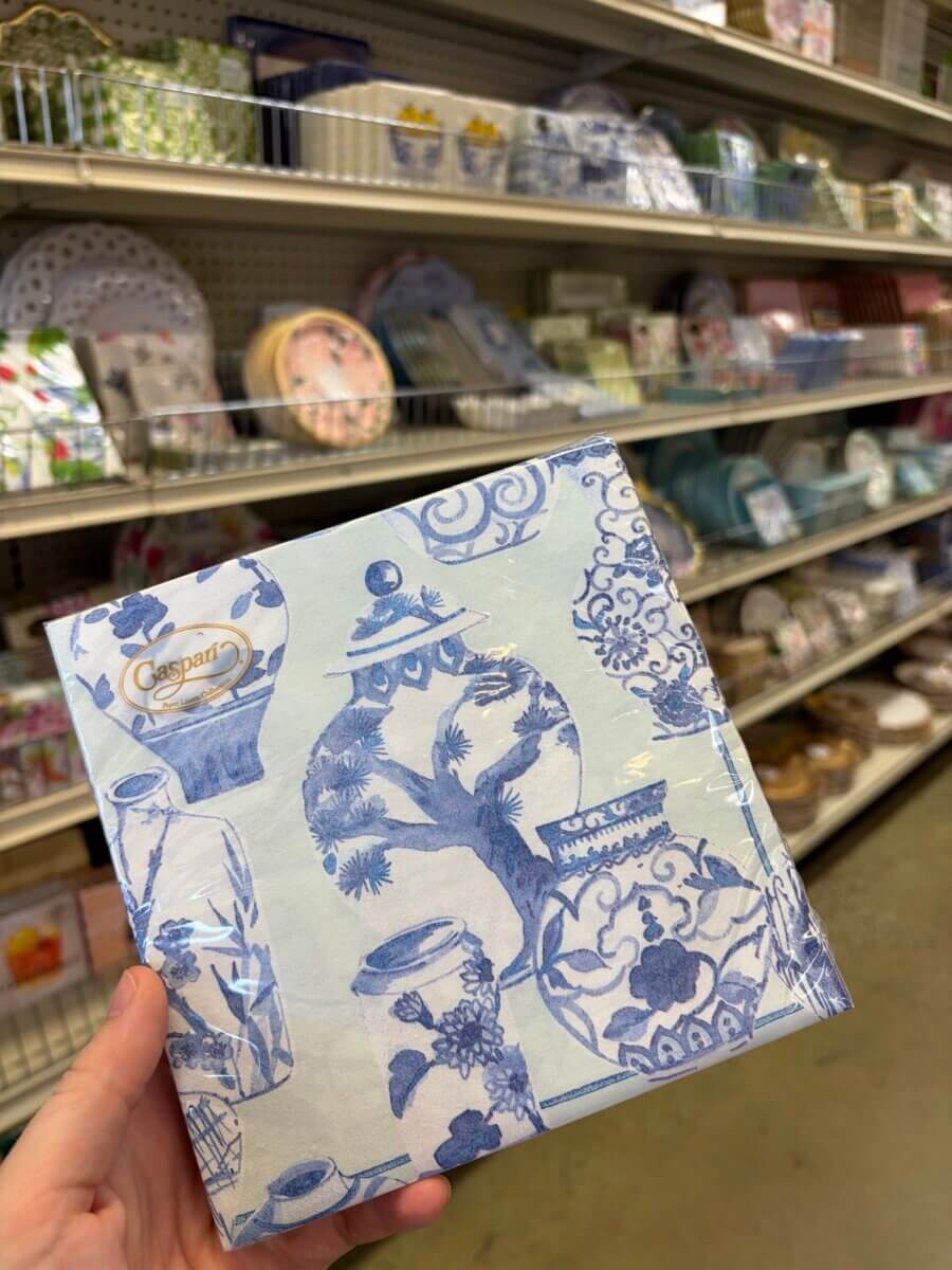 15 Birmingham FINDS for August: Breezy Fashion, Entertaining & More - 3 A hand holds blue and white decorative napkins with a vase pattern in front of store shelves displaying various tableware and home decor FINDS.