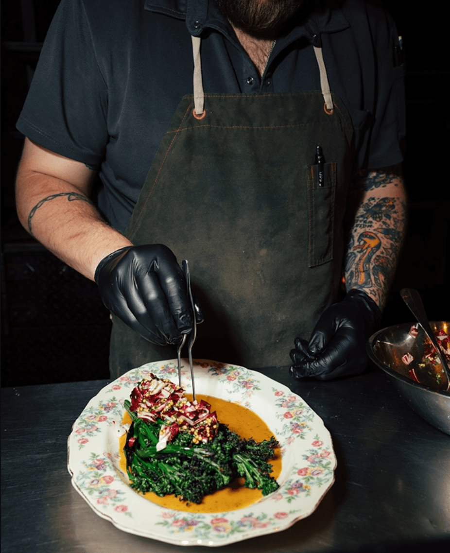 A chef in black gloves plates a dish with greens and sauce on a floral-patterned plate, wearing a dark shirt and apron, with tattooed arms visible—bringing artistry to the table for Birmingham Restaurant Week.