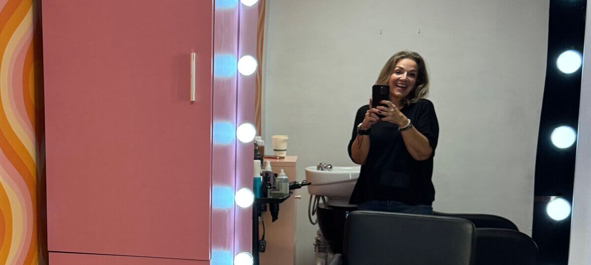 A woman stands and smiles while taking a selfie in front of a mirror with round lights at Sanctum Salon, next to a pink cabinet.