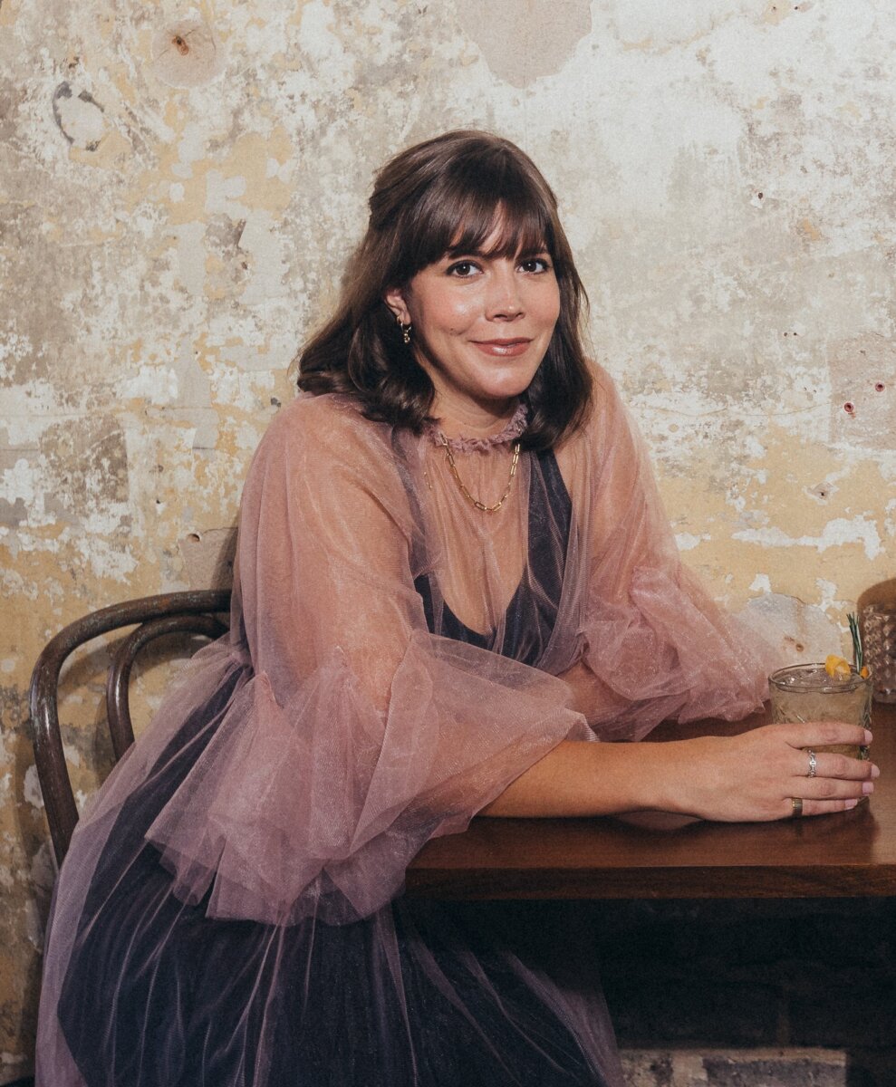 Ali Randall in a sheer pink dress sits at a wooden table against a textured wall, holding a drink with a garnish and looking at the camera, set behind one of the prettiest bars in BHAM.