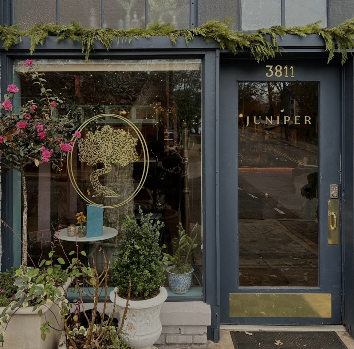 Juniper storefront with large window featuring a tree logo, potted plants outside, and "JUNIPER" and address "3811" displayed on the door. Greenery is draped above the entrance, making it one of BHAM's prettiest bars—even more so when you know she's behind such a charming design.