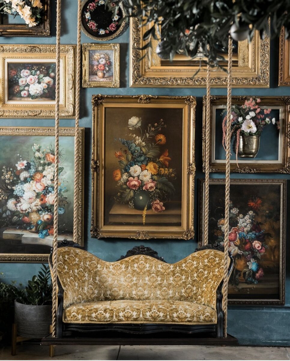 Antique floral paintings in gold frames cover a blue wall behind a vintage yellow upholstered swing sofa with rope supports.