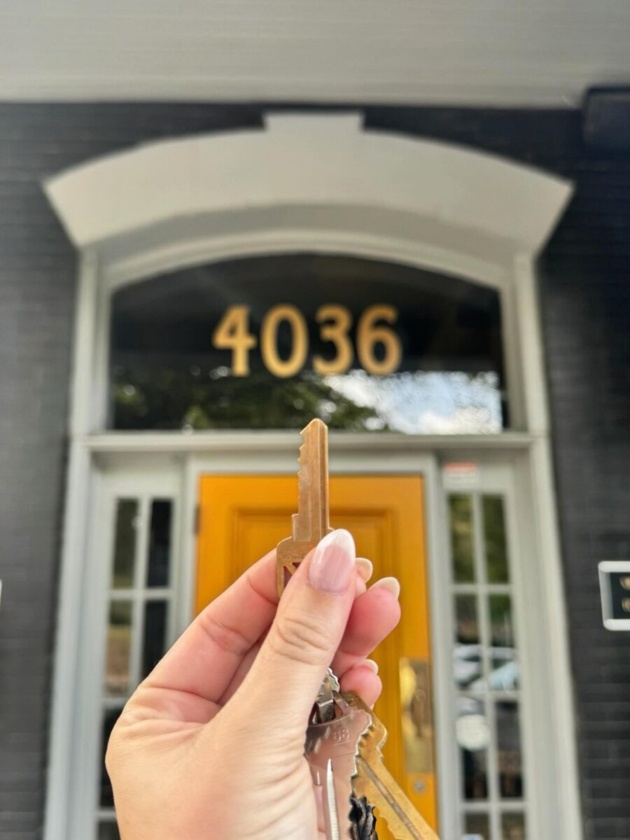 A hand holding up a set of keys in front of a yellow door with the address number 4036 displayed above.