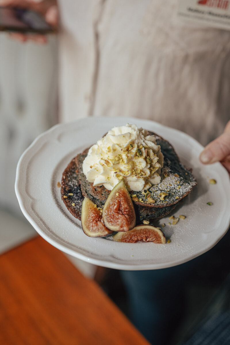 A person holds a plate with pancakes topped with whipped cream, powdered sugar, crushed pistachios, and fresh fig slices.