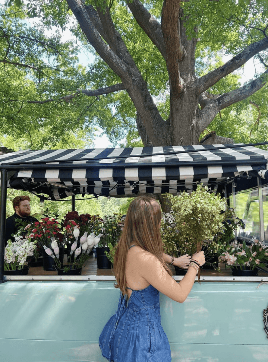 5 U-Pick Flower Farms Near Birmingham - 5 A woman in a blue dress arranges flowers at an outdoor flower stand with a black-and-white striped canopy, under a large tree at one of Birminghamβs charming U-Pick Flower Farms.