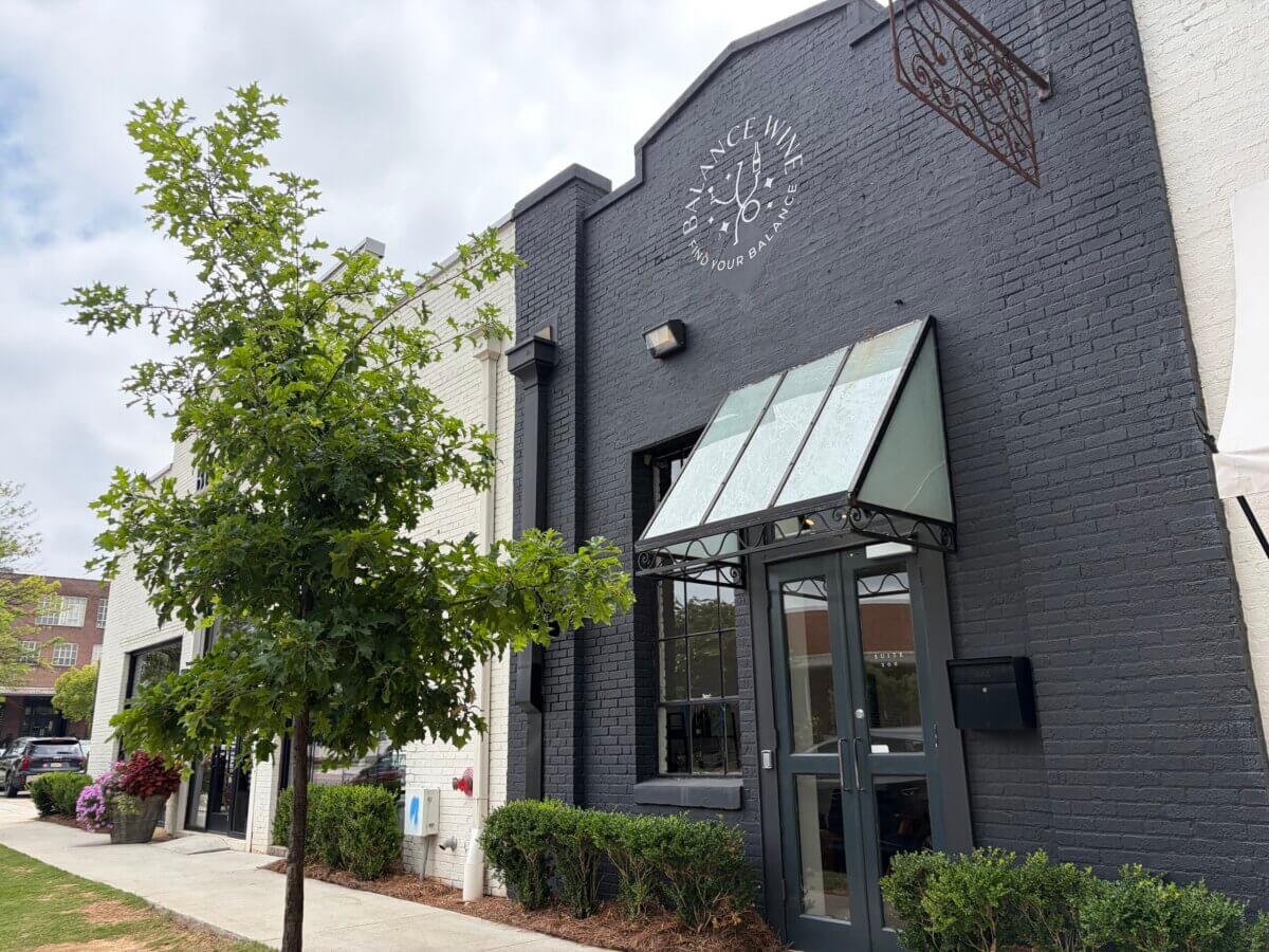 Your New Favorite Wine Destination Just Opened in Pepper Place - 4 Exterior of a brick restaurant at Pepper Place with a large glass awning, gray and white walls, a sign above, and a small tree and shrubs along the sidewalk.
