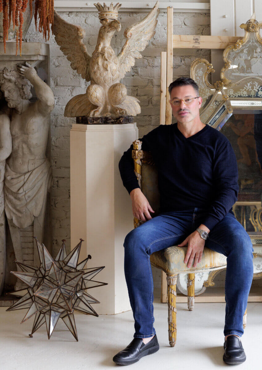 A man in a black sweater and jeans sits on an ornate chair in a room decorated with classical statues, a large mirror, and a sculpted eagle—an elegant space curated by Deke Rogers as part of his Collected for the Home antiquing adventures.