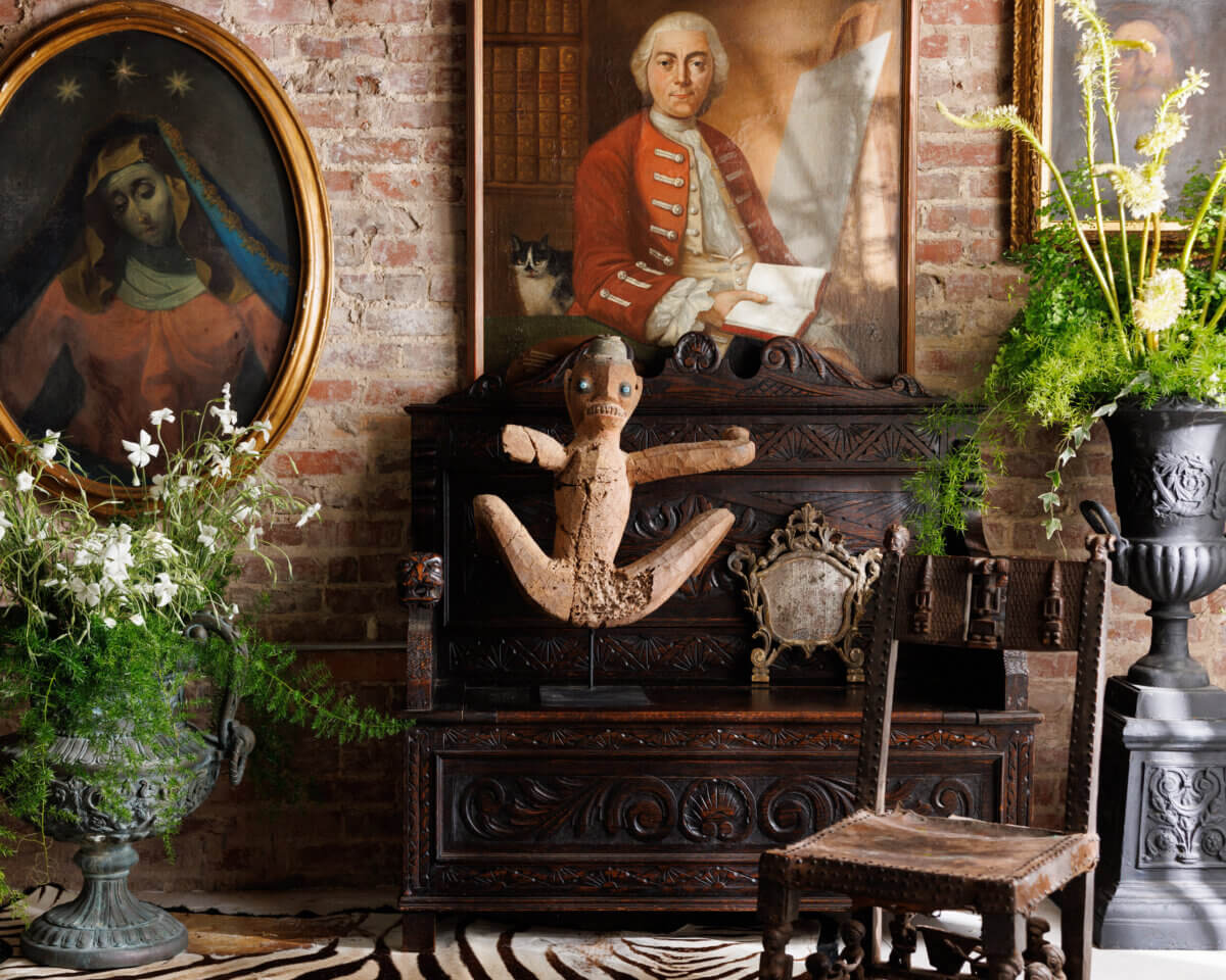 Eclectic interior with ornate wood furniture, a doll sculpture, two framed portraits, lush green plants, and a zebra-patterned rug on a brick wall background.