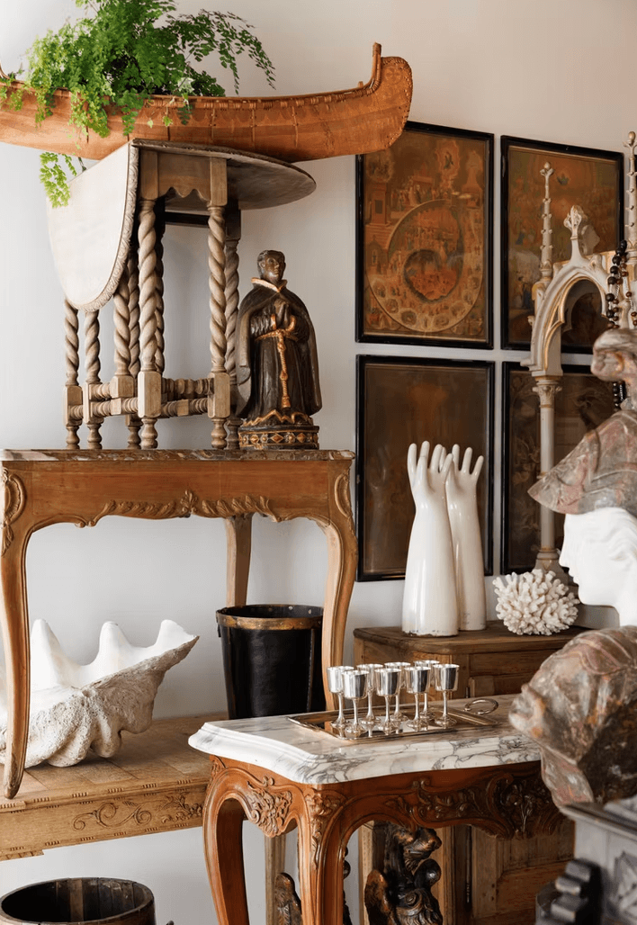 An eclectic room features ornate tables, framed art, a plant, statues, white hand sculptures, a large shell, and various decorative objects arranged closely together.