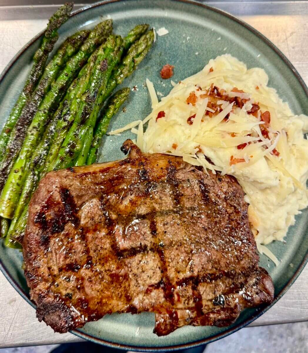 11 New Birmingham Restaurants to Try This Month - 2 Grilled steak with crosshatch marks, roasted asparagus, and mashed potatoes topped with shredded cheese and bacon bits on a green plate.