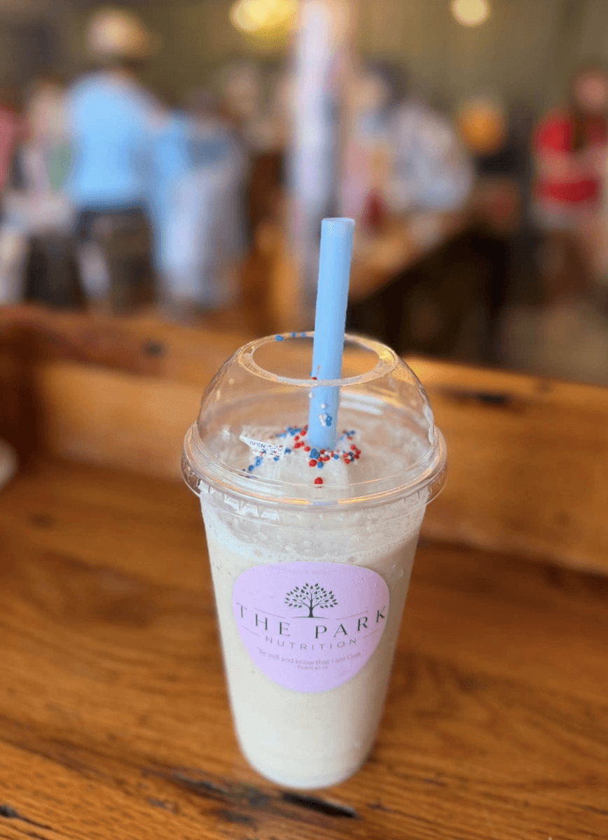 11 New Birmingham Restaurants to Try This Month - 11 A plastic cup with a dome lid and blue straw holds a creamy drink topped with sprinkles. The label reads "The Park Nutrition." Perfect for fans of dining at Birmingham restaurants, the cup sits on a wooden table in a busy indoor setting.