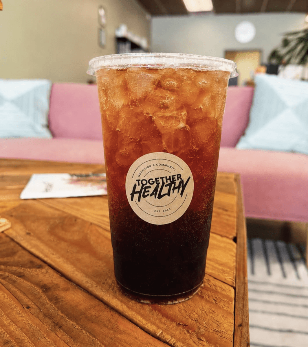 11 New Birmingham Restaurants to Try This Month - 5 A plastic cup of iced tea with a "Together Healthy" logo sits on a wooden table in front of a pink couch.