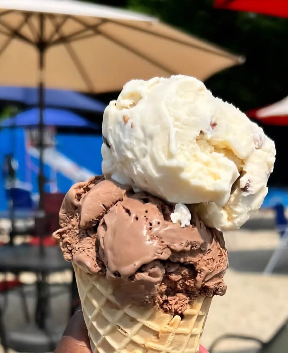 11 New Birmingham Restaurants to Try This Month - 6 A hand holds a waffle cone with two scoops of ice cream, one chocolate and one white, outdoors near tables, chairs, and umbrellas.