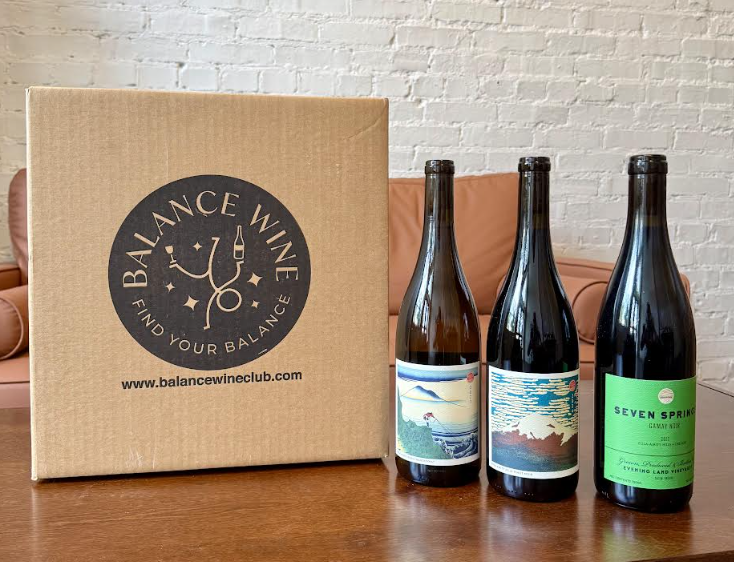 Your New Favorite Wine Destination Just Opened in Pepper Place - 2 Three bottles of Balance Wine stand beside a Balance Wine Club box on a wooden table, with a sofa and white brick wall in the background, evoking the welcoming charm of Birmingham.