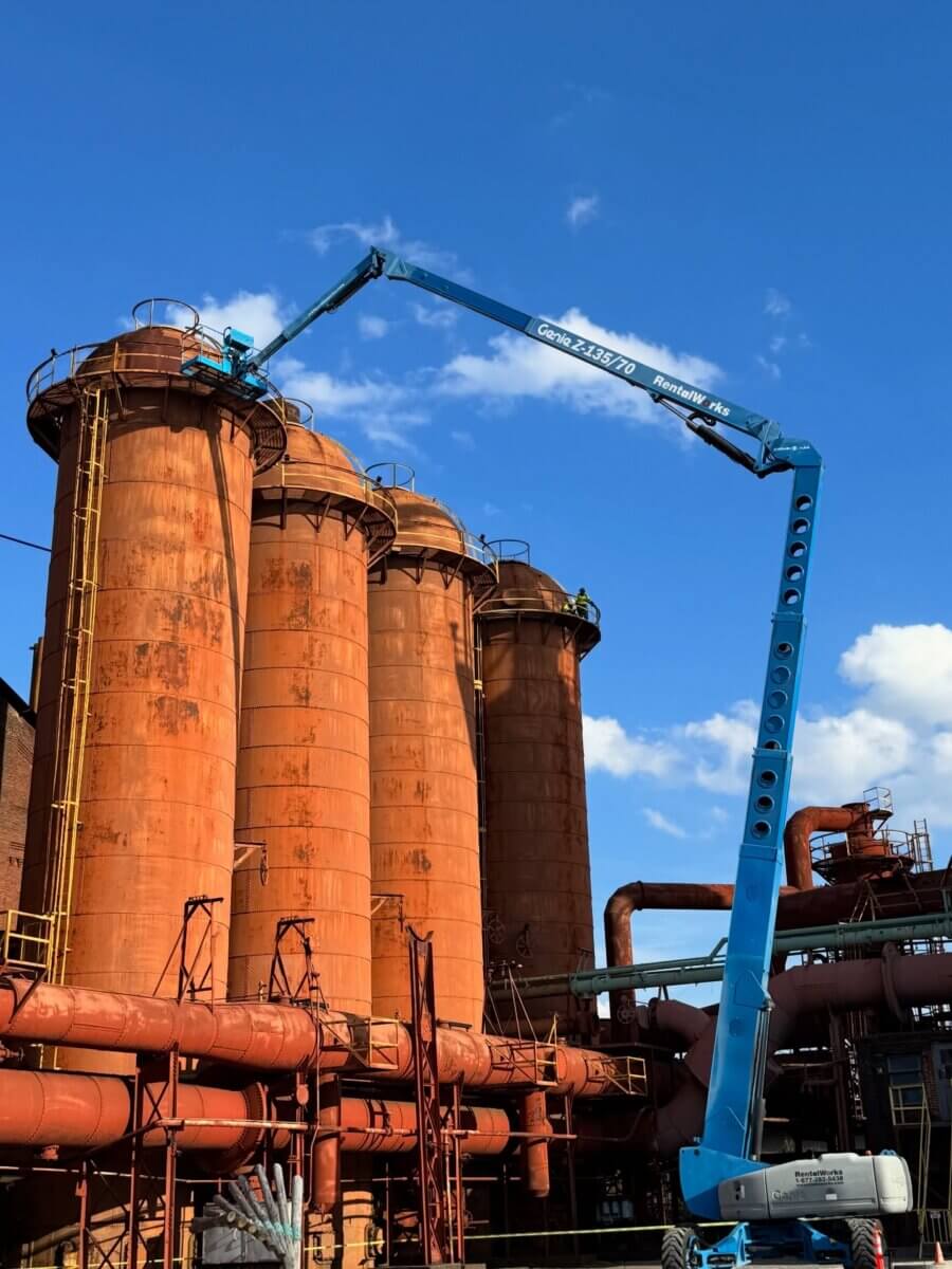 A blue boom lift extends to the top of large rusted industrial silos and pipes at Sloss Furnaces, reaching skyward under a clear blue sky with some clouds.