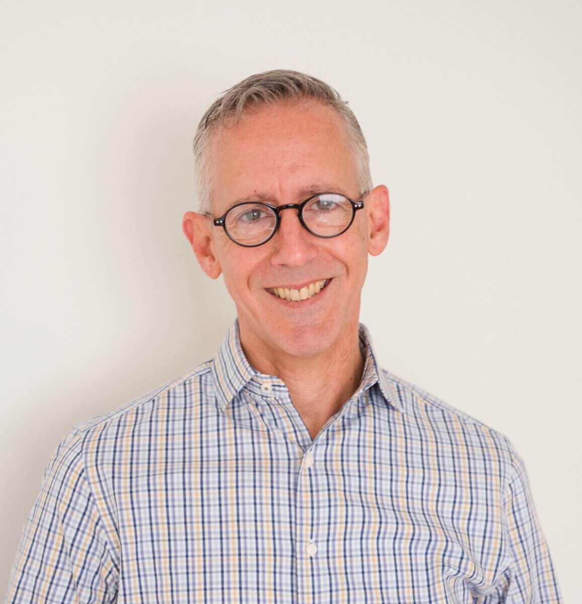 Smiling man with short gray hair and glasses wearing a blue and white plaid shirt, standing in front of a plain white background.