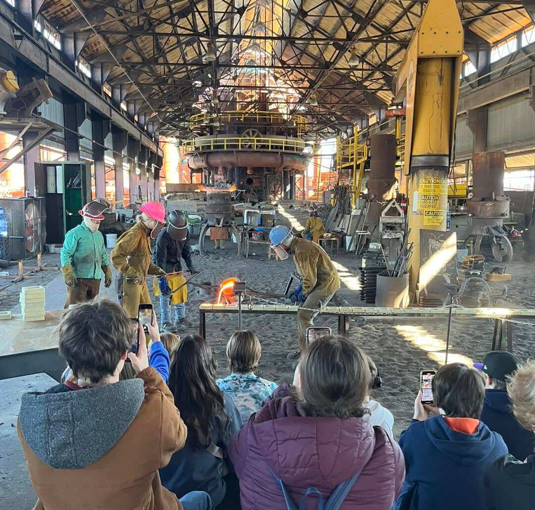 People watch as workers in protective gear pour molten metal in a large industrial foundry at Sloss Furnaces; several audience members scoop up photos with their phones, eager to capture the fiery spectacle.