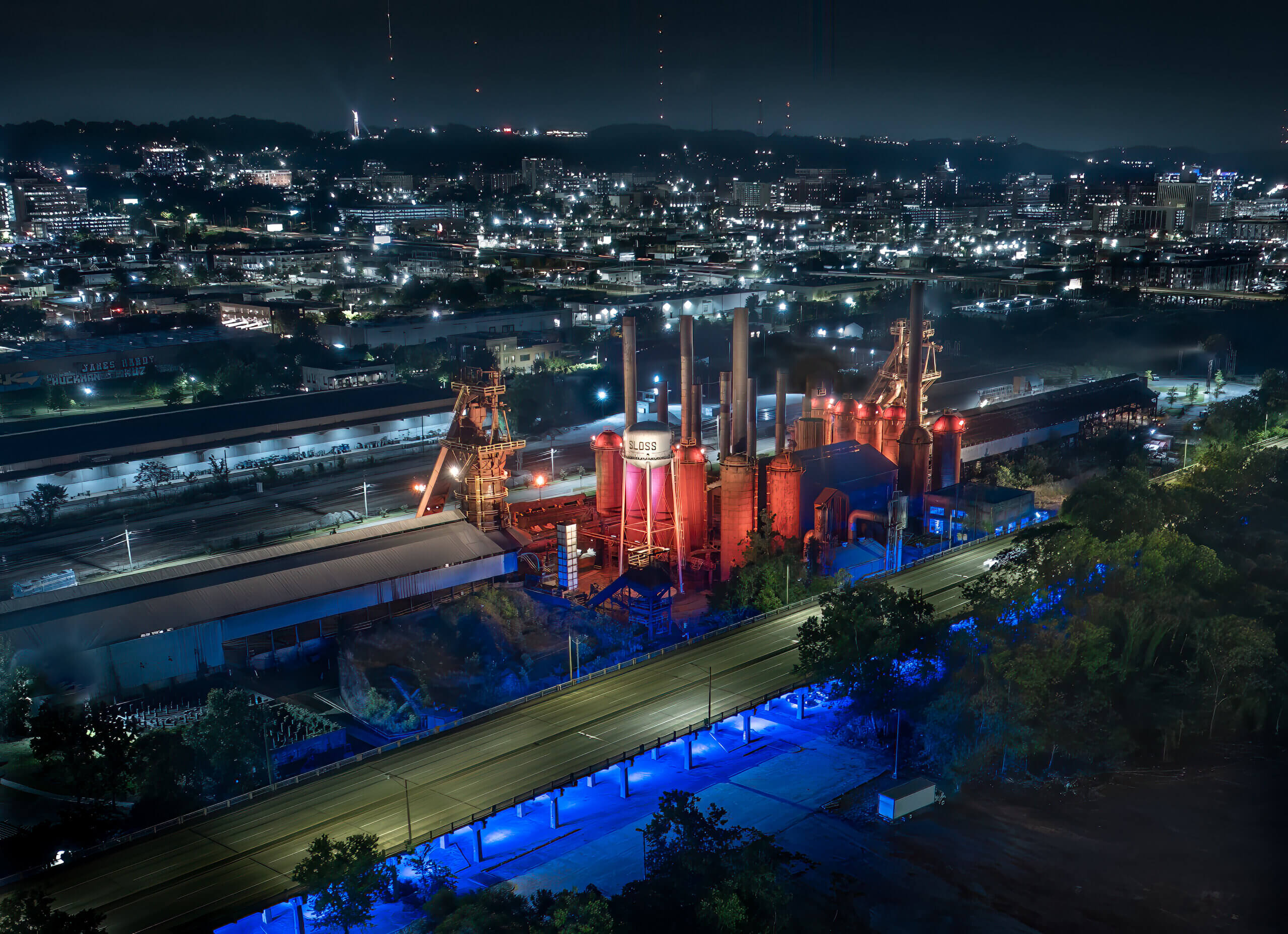 Aerial night view of Sloss Furnaces, an industrial plant with red-lit smokestacks and surrounding city lights, bordered by a brightly illuminated blue road in the foreground—a scene that hints at a major makeover on the horizon.