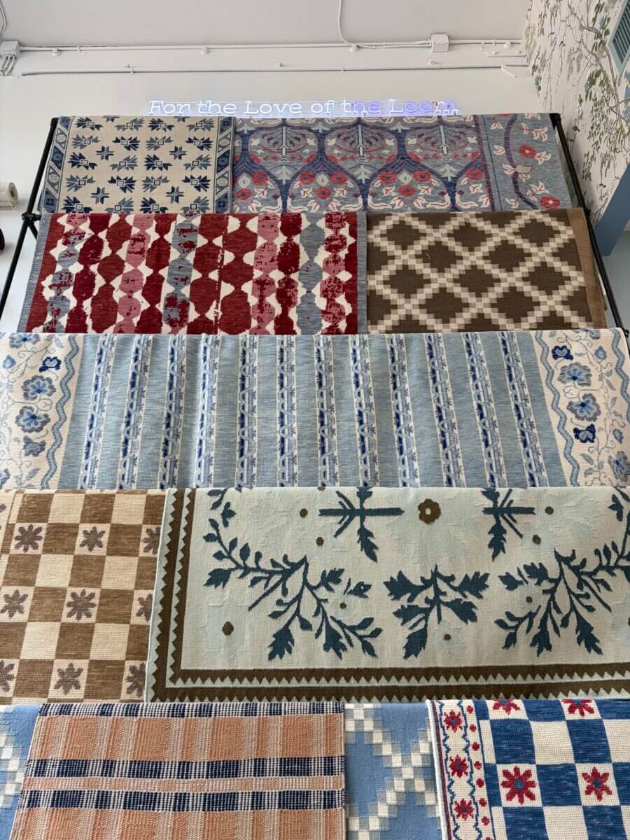 This Award-Winning BHAM Rug Company Launched a New Collection Celebrating America’s 250th Year - 2 A display of ten patterned rugs from Birmingham Rug Company's new collection, arranged in two rows against a wall with a blue neon sign, celebrates America’s 250th Year with vibrant colors and unique designs.