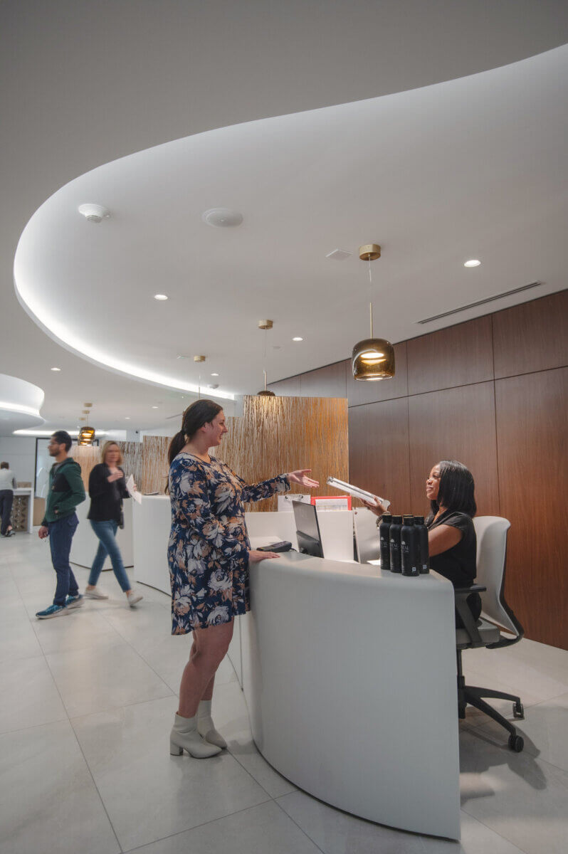 A woman in a floral dress speaks with a receptionist at a modern, white curved front desk, possibly inquiring about tanning bed risks; three people walk by in the background.