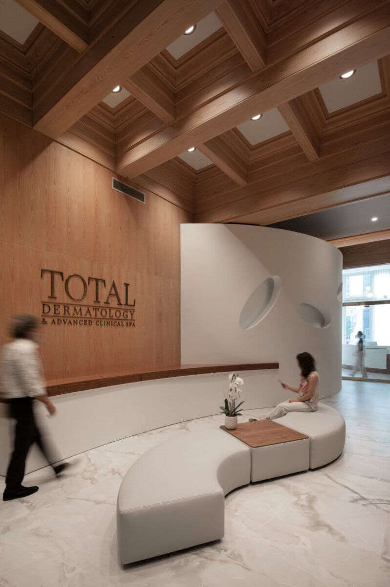 Modern dermatology clinic lobby with wood and white decor, curved seating, an orchid plant, two people, and the "Total Dermatology & Advanced Clinical Spa" sign on the wall—a welcoming space where experts educate patients about skin health and tanning bed risks.