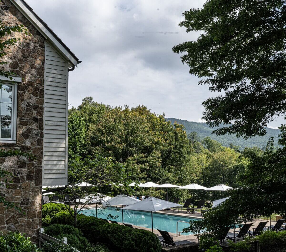 8 Luxurious, Family-Friendly Resorts in the South - 9 Outdoor pool area with lounge chairs and white umbrellas, bordered by greenery and trees, next to a stone building; mountains visible in the background under a cloudy sky—an inviting setting perfect for Southern families seeking relaxing escapes at family-friendly resorts.