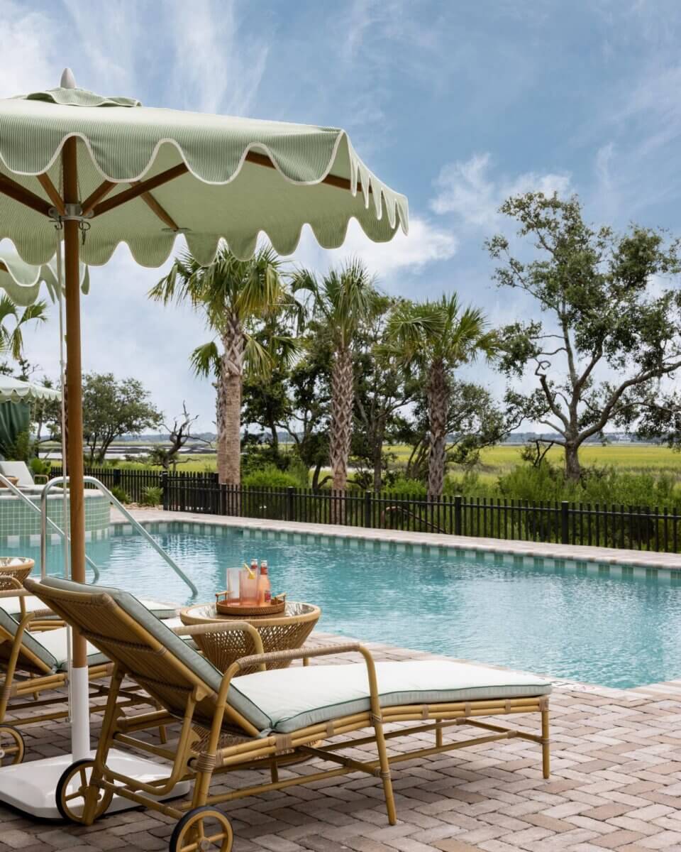 8 Luxurious, Family-Friendly Resorts in the South - 6 Two lounge chairs with cushions under a scalloped umbrella by a clear swimming pool, with palm trees and a grassy landscape in the background. A small table holds drinks and towels, setting the perfect scene for Southern families seeking relaxation at family-friendly resorts.