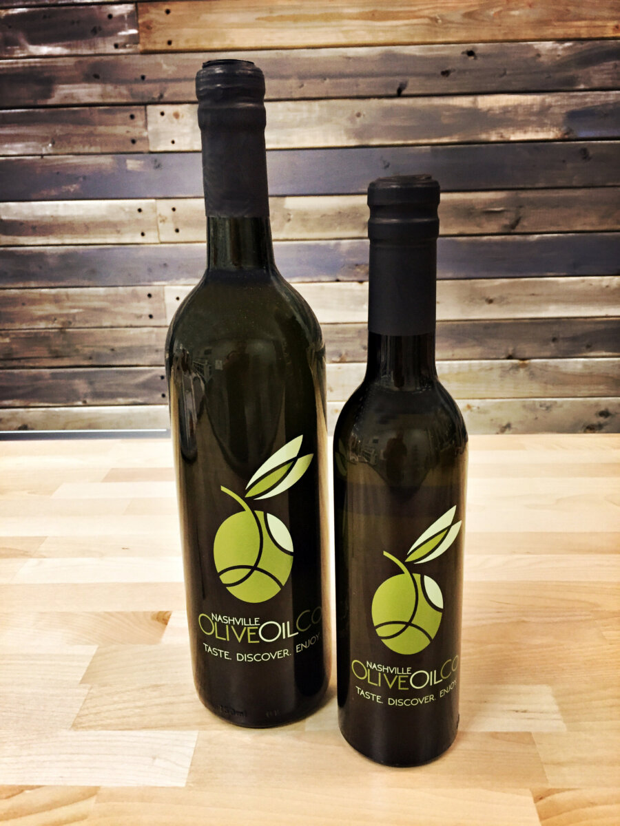 Two dark glass bottles labeled "Nashville Olive Oil Co." with green olive branch logos stand on a light wooden surface against a wooden plank background.